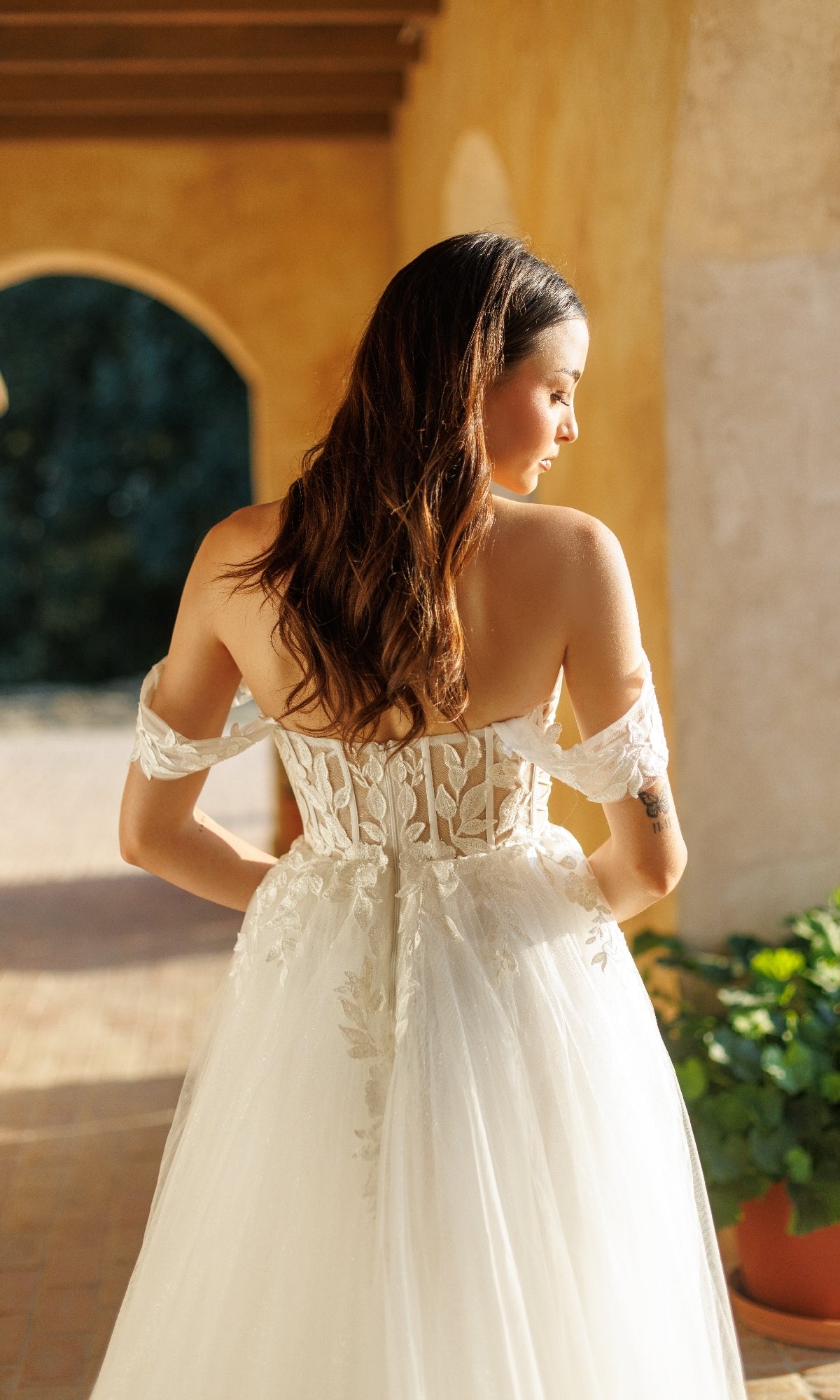 A woman in the Tina Holly TW480 Long Formal Dress, featuring an off-shoulder lace-embellished corset bodice, stands outdoors with her back to the camera as warm sunlight highlights its romantic details.
